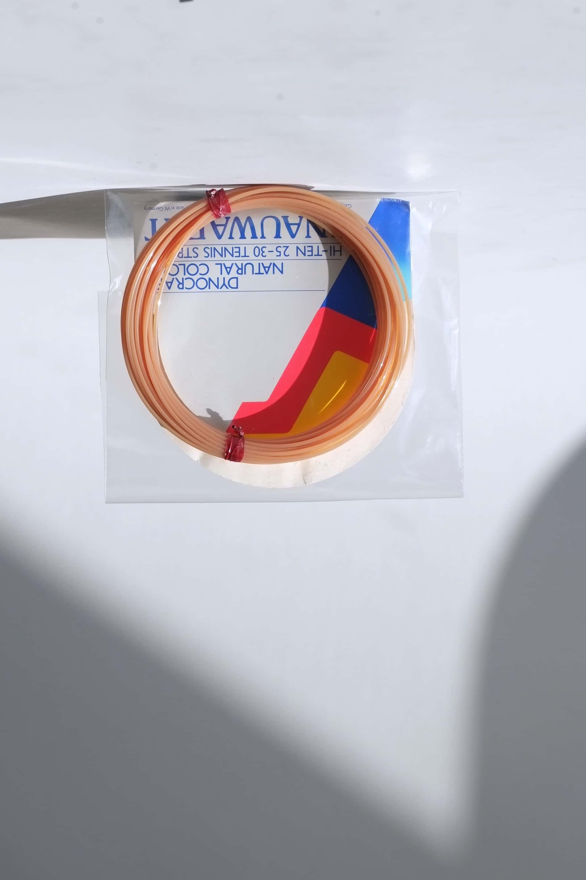 coiled vintage Snauwaert Dynocraft Hi-Ten 25-30 tennis strings in their original grey, yellow, and red and blue retail sleeve packaging.