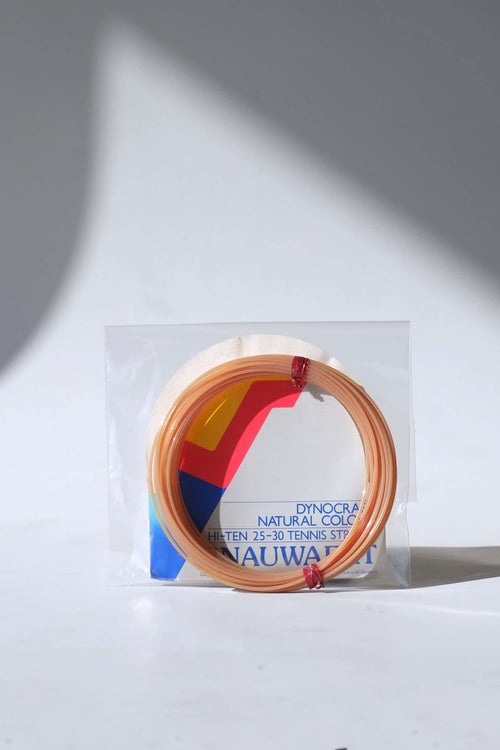 coiled vintage Snauwaert Dynocraft Hi-Ten 25-30 tennis strings in their original grey, yellow, and red and blue retail sleeve packaging.