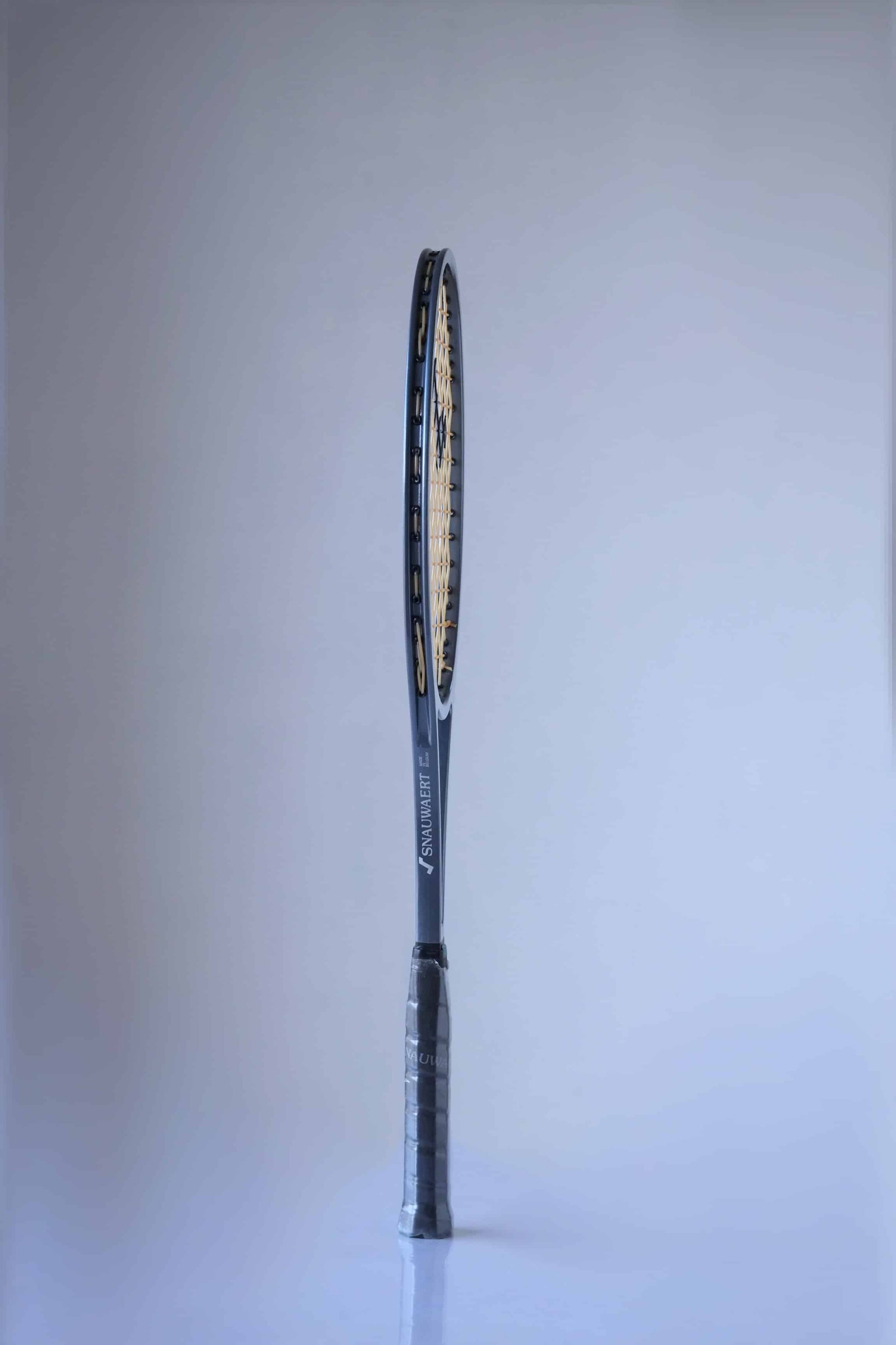 Side view of a vintage Snauwaert Hi-Ten25 tennis racquet. The frame is grey with the logo 'Snauwaert' printed in light grey, along the side. The racquet handle is black and wrapped in plastic.