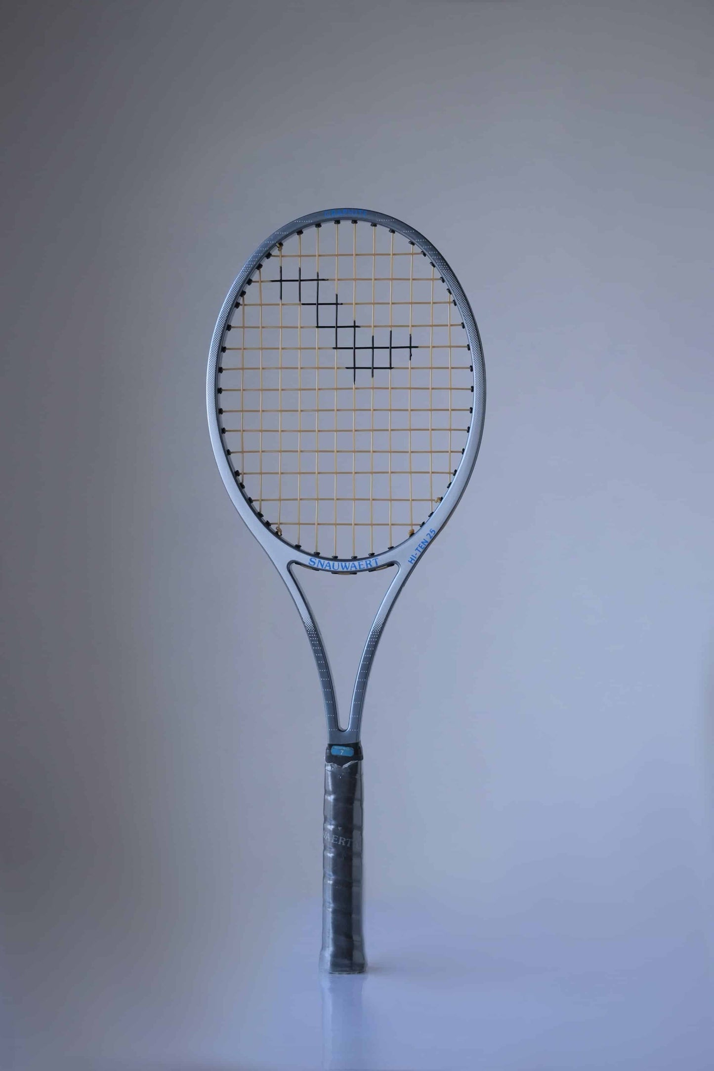 Full view of a Snauwaert Hi-Ten 25 tennis racket, showing its grey frame, yellow strings with a black pattern, and wrapped handle. The Snauwaert logo is visible on the throat of the racket.