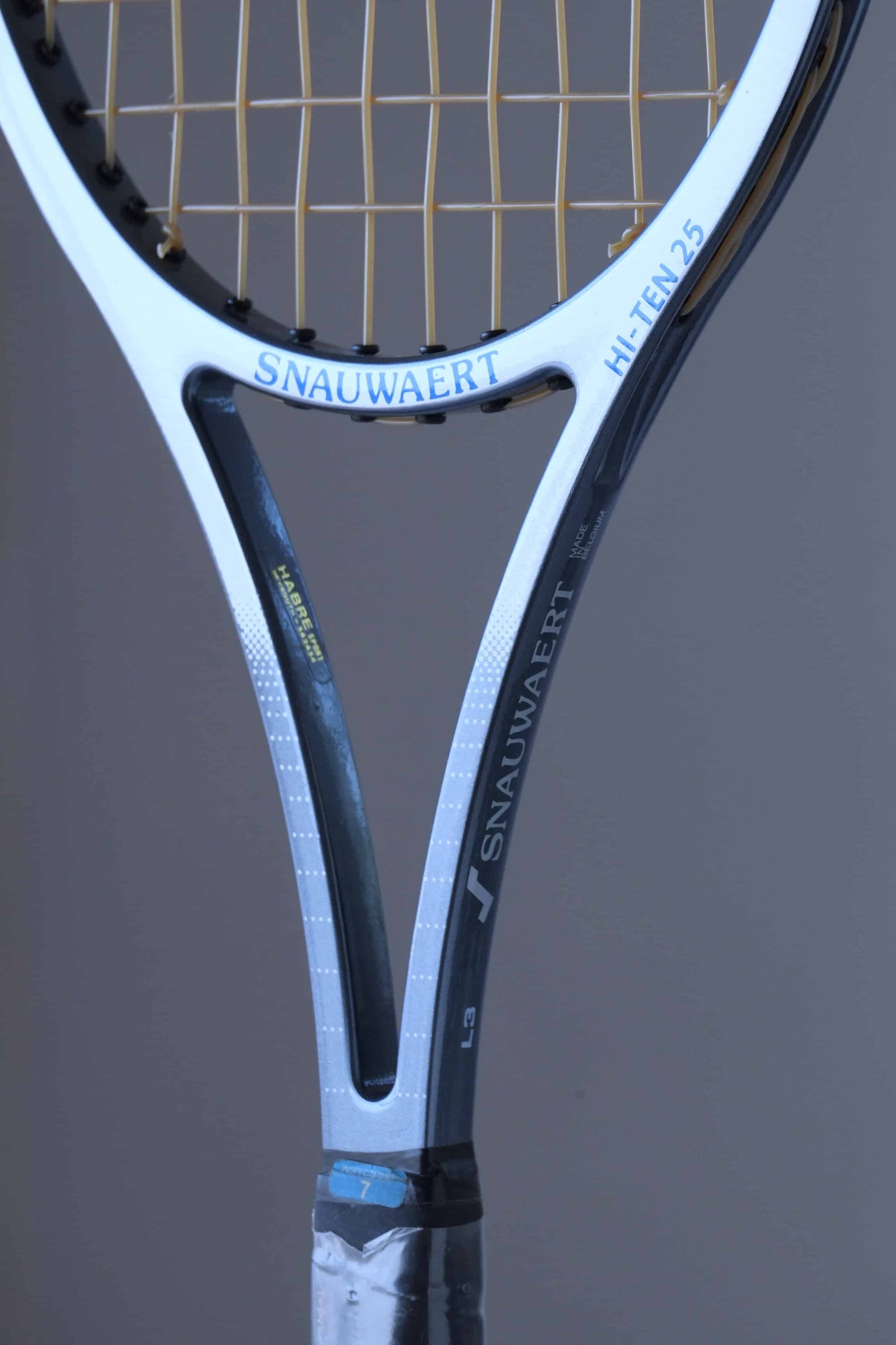 Close-up view of a Snauwaert Hi-Ten 25 tennis racket throat. The logo 'Snauwaert' is printed in blue, with 'Hi-Ten 25' marked in white along the side. The frame is a combination of white and grey with the strings attached to the head, and part of the wrapped handle is visible.