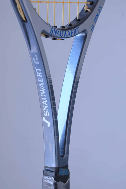 Close-up view of a Snauwaert Hi-Ten 25 tennis racket throat. The logo 'Snauwaert' is printed in blue, with 'Hi-Ten 25' marked in white along the side. The frame is a combination of white and grey with the strings attached to the head, and part of the wrapped handle is visible.