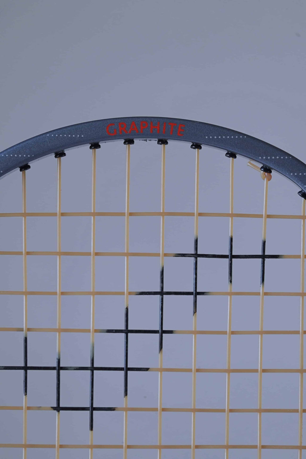 Close-up of the top section of a Snauwaert Hi-Ten 30 tennis racket, showing the word 'GRAPHITE' in red on the grey frame, with yellow strings and a black patterned string design.