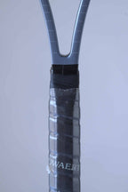 Close up of the racquet handle of a vintage Snauwaert Hi-Ten 30 tennis racket, showing part of the word 'SNAUWAERT' in grey. The handle is wrapped with black overgrip tape and covered with plastic.
