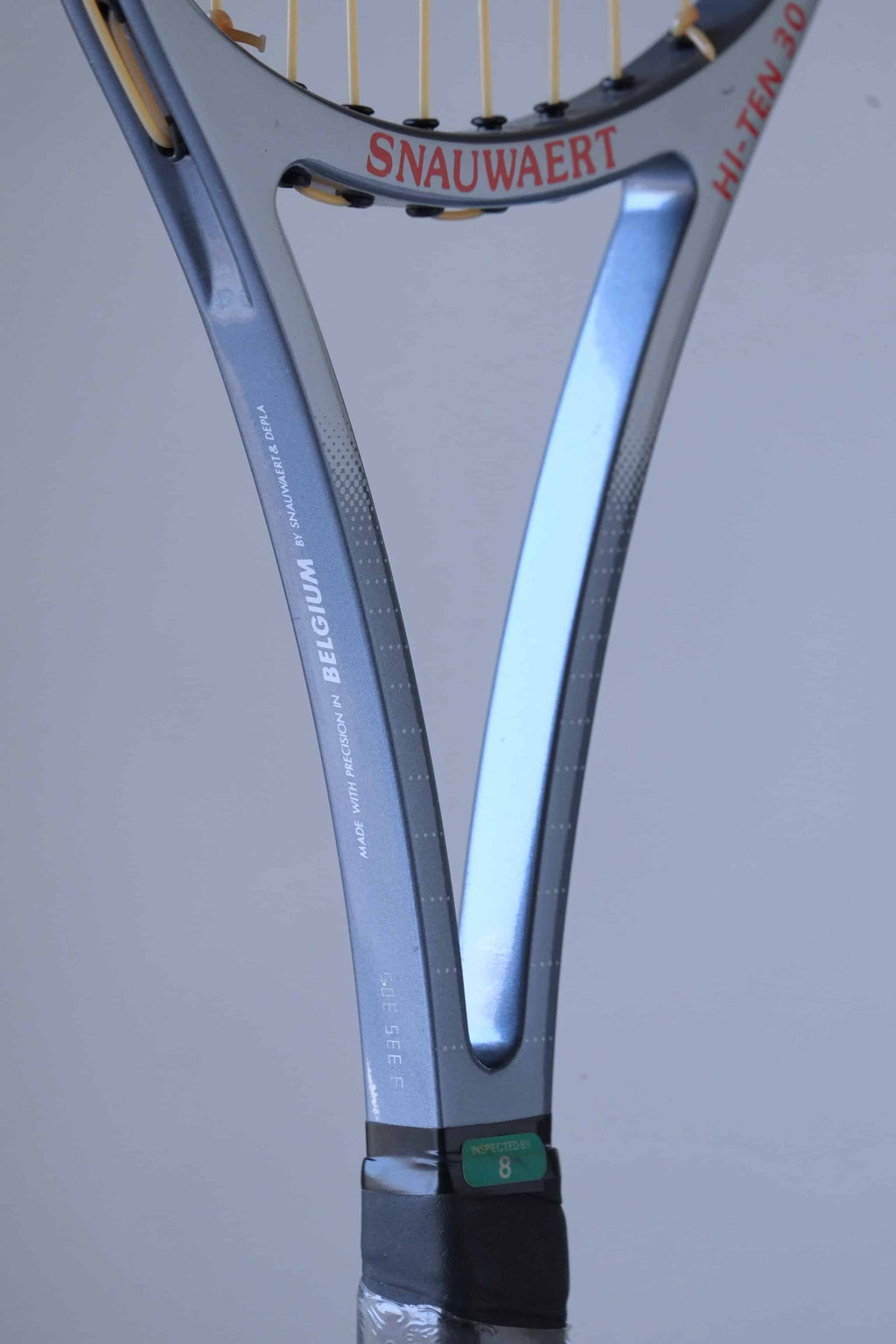 Close-up of a Snauwaert tennis racket handle. The handle is blue and black with a textured grip. The word "SNAUWAERT" is written in white on the handle, and the number "8" is visible below it. The racket is made in Belgium.