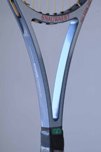 Close-up of a Snauwaert tennis racket handle. The handle is blue and black with a textured grip. The word "SNAUWAERT" is written in white on the handle, and the number "8" is visible below it. The racket is made in Belgium.