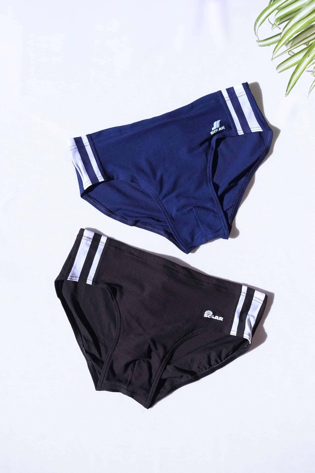 Two pairs of vintage SOLAR 70s swim briefs in navy and black with white double stripe detailing and printed logo, laid flat on white background.