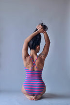 Solar vintage 80s pink and blue block stripe spaghetti strap leotard worn on a female model — wide equal-weight horizontal stripes, cross-back spaghetti straps, open back, hands gathering hair overhead, seated pose, rear view