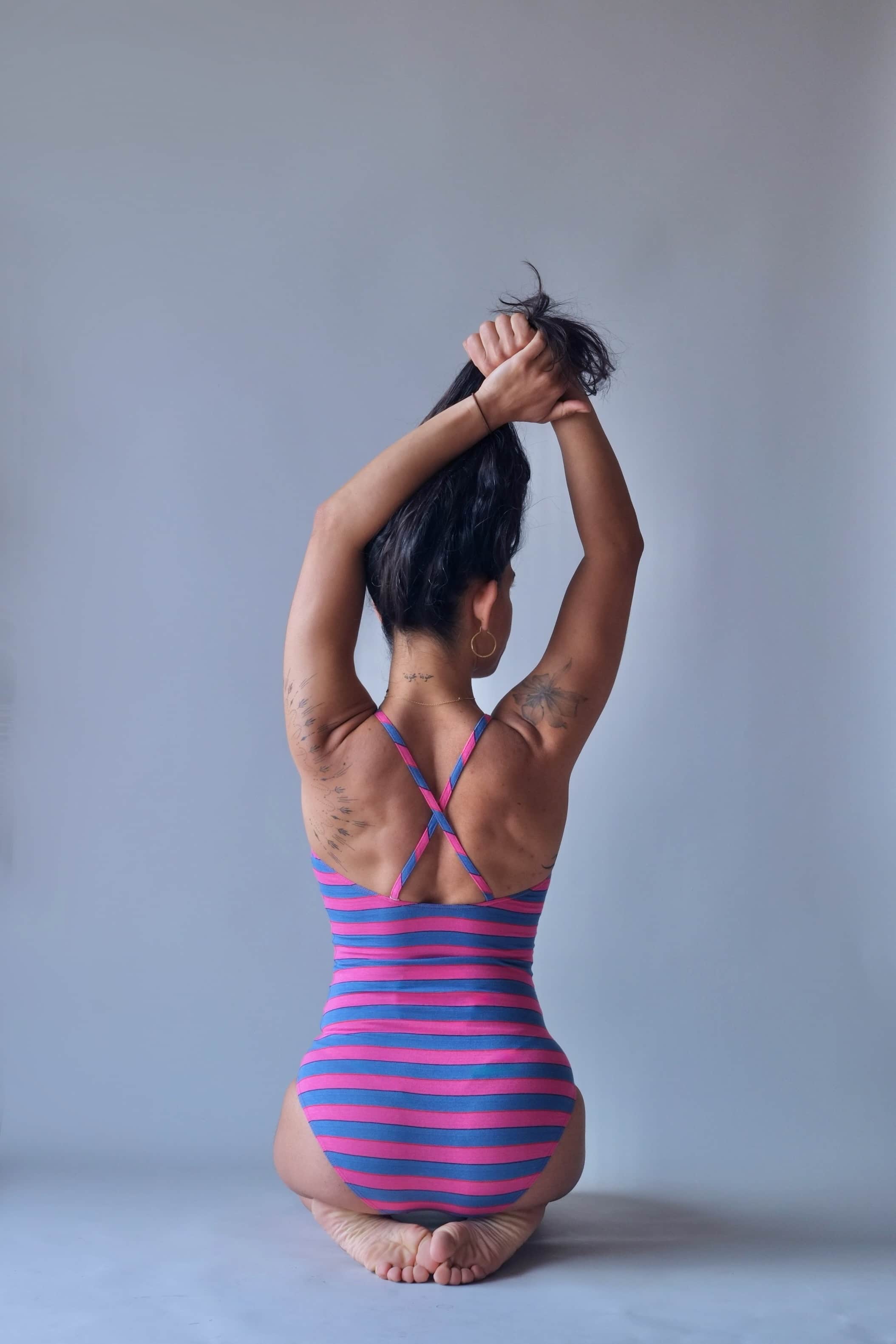 Solar vintage 80s pink and blue block stripe spaghetti strap leotard worn on a female model — wide equal-weight horizontal stripes, cross-back spaghetti straps, open back, hands gathering hair overhead, seated pose, rear view