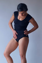 Solar vintage 80s solid black cap sleeve leotard worn on a female model — scoop neckline, fitted cap sleeves, high cut leg, dynamic standing pose, front view