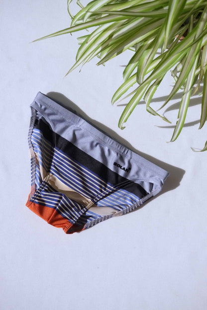 A pair of SOLAR brand vintage 80s men's swim briefs with multi-colored horizontal stripes in grey, black, and orange tones, laid flat on a white background.