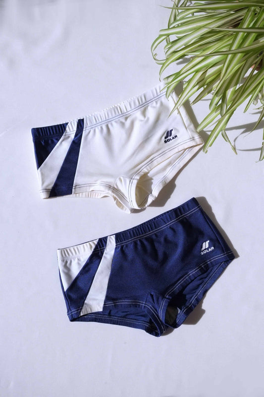 Two pairs of SOLAR brand vintage 80s men's square-cut swim briefs: one off-white with navy diagonal block, the other navy with white diagonal block, laid flat on a white background..