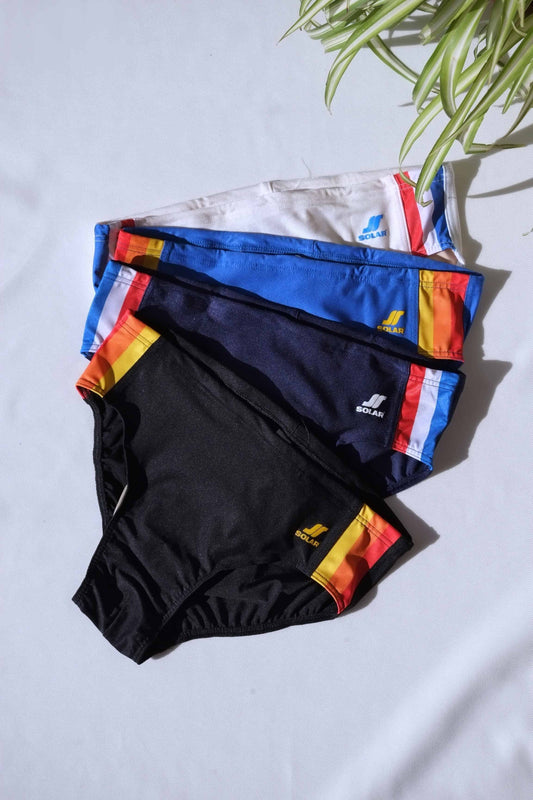 Four pairs of SOLAR brand vintage 80s men's swim briefs in white, blue, navy, and black, each with multi-colored striped side panels, laid flat under sunlight.