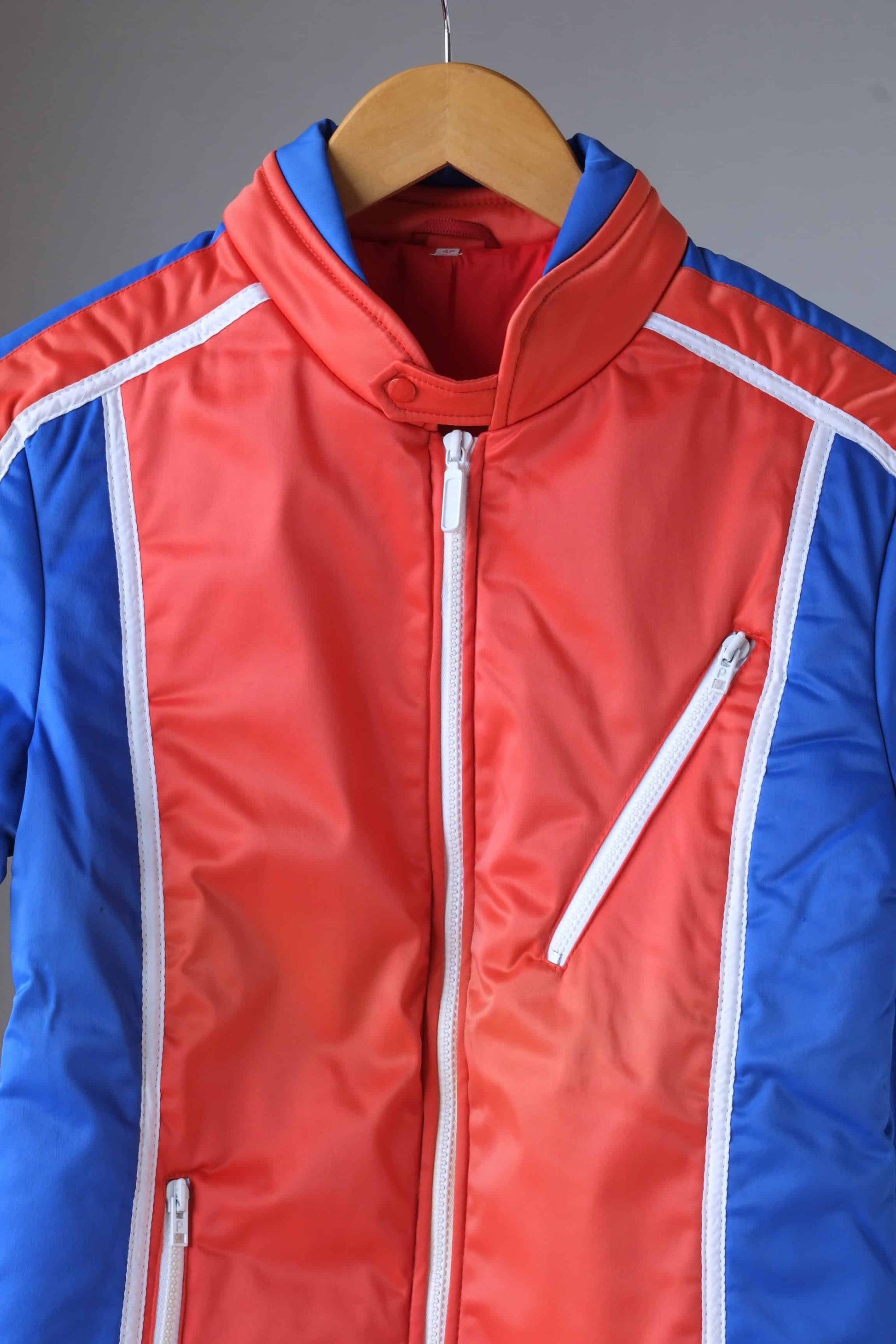 Close up view of a vintage Sportalm ski jacket. The jacket is red and blue with white piping and a zipper closure, displayed on a hanger.