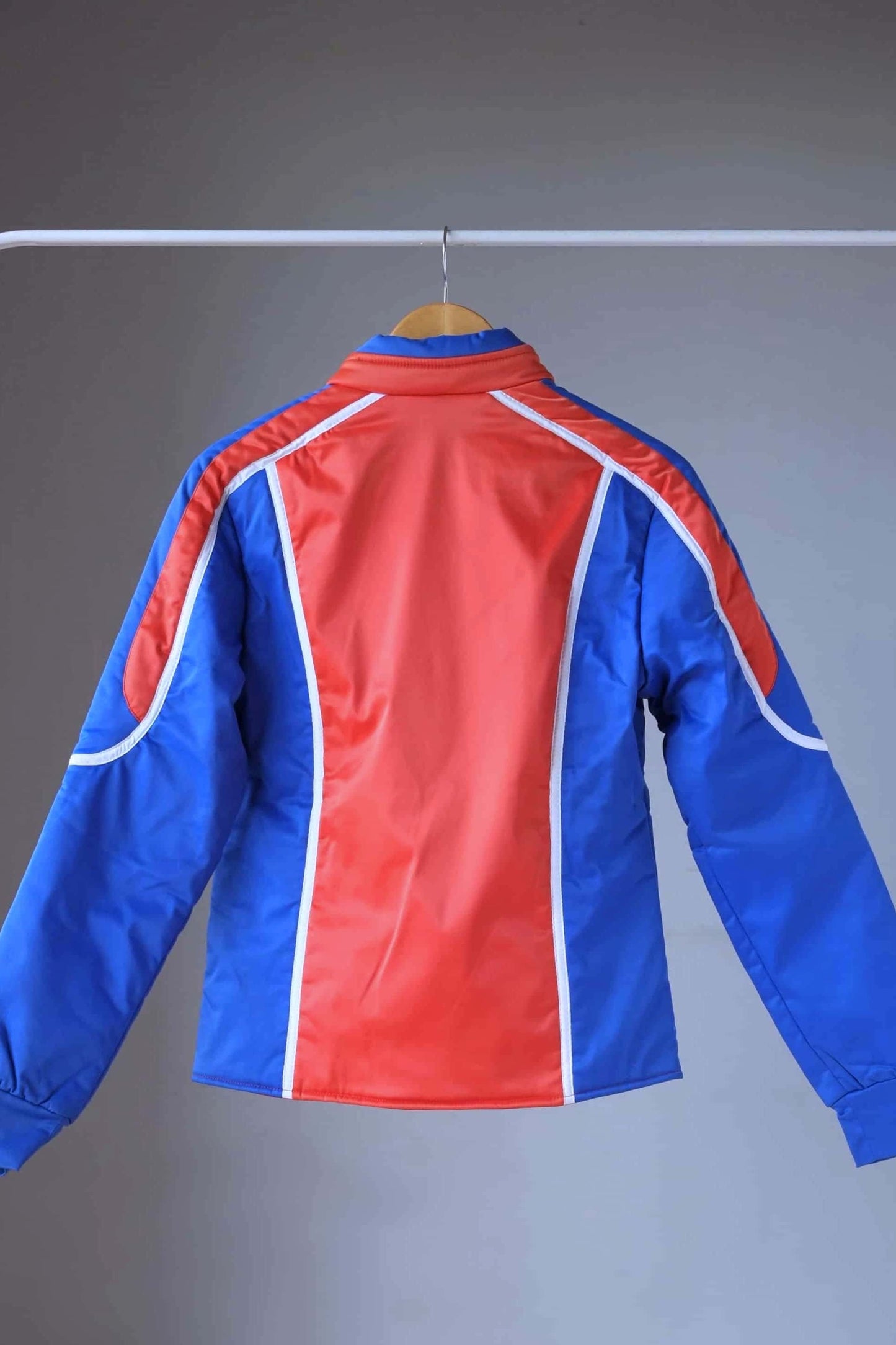 Back view of a vintage Sportalm ski jacket. The jacket is red and blue with white piping and a zipper closure, displayed on a hanger.