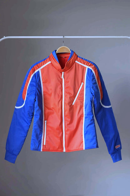 Front view of a vintage Sportalm ski jacket. The jacket is red and blue with white piping and a zipper closure, displayed on a hanger.