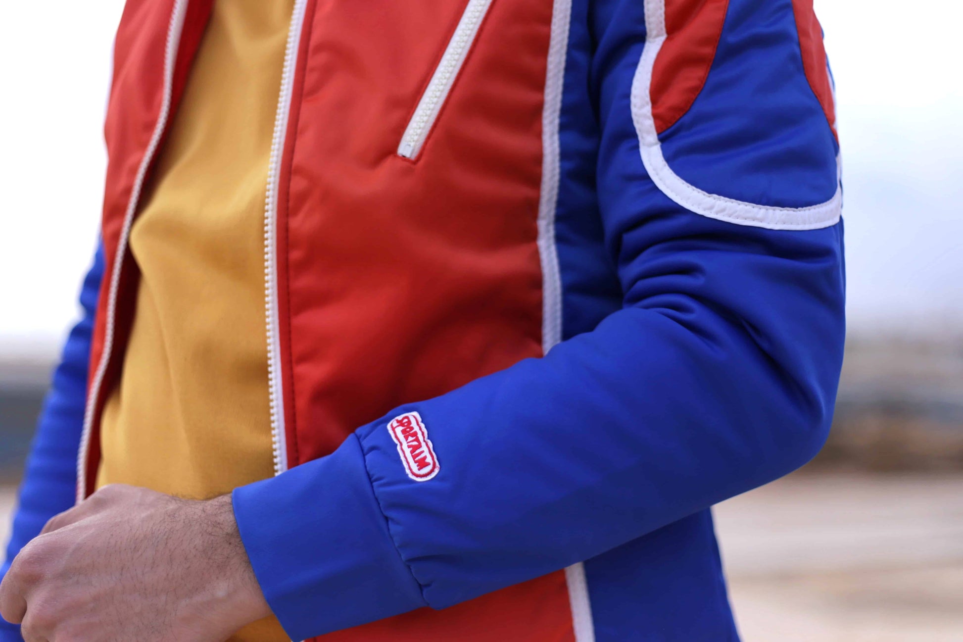 Close view of a vintage Sportalm ski jacket. The jacket is red and blue with white piping and a zipper closure. It is worn by a model over a mustard yellow top.