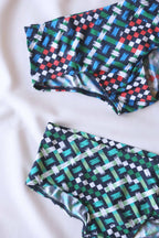 Close up of Two pairs of men's swim briefs in a geometric pattern of blue,  green, white, red, and black squares and lines, laid out on a white surface