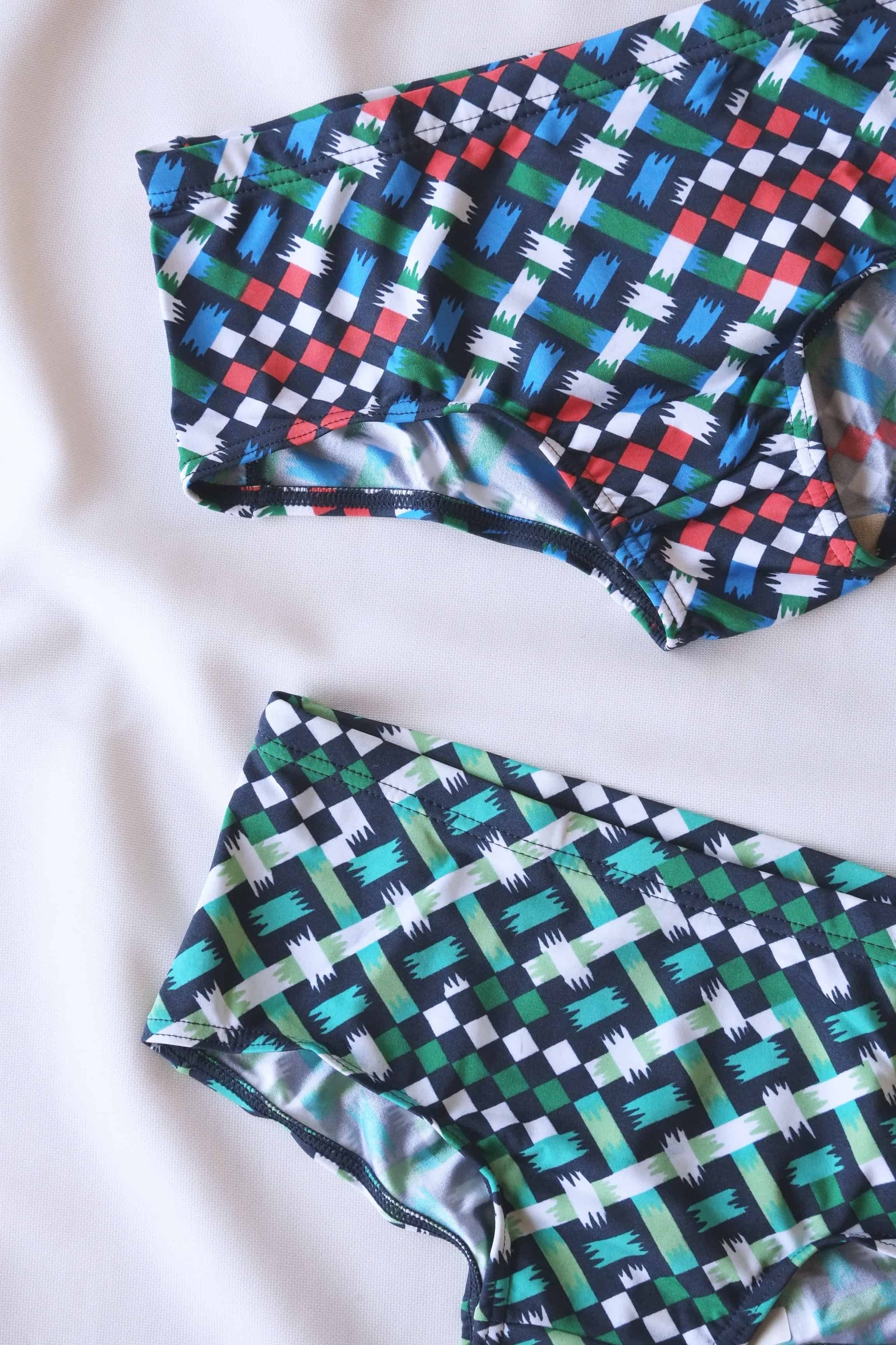 Close up of Two pairs of men's swim briefs in a geometric pattern of blue,  green, white, red, and black squares and lines, laid out on a white surface