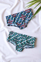 Two pairs of men's swim briefs in a geometric pattern of blue, green, white, red, and black squares and lines, laid out on a white surface with a green plant in the background.