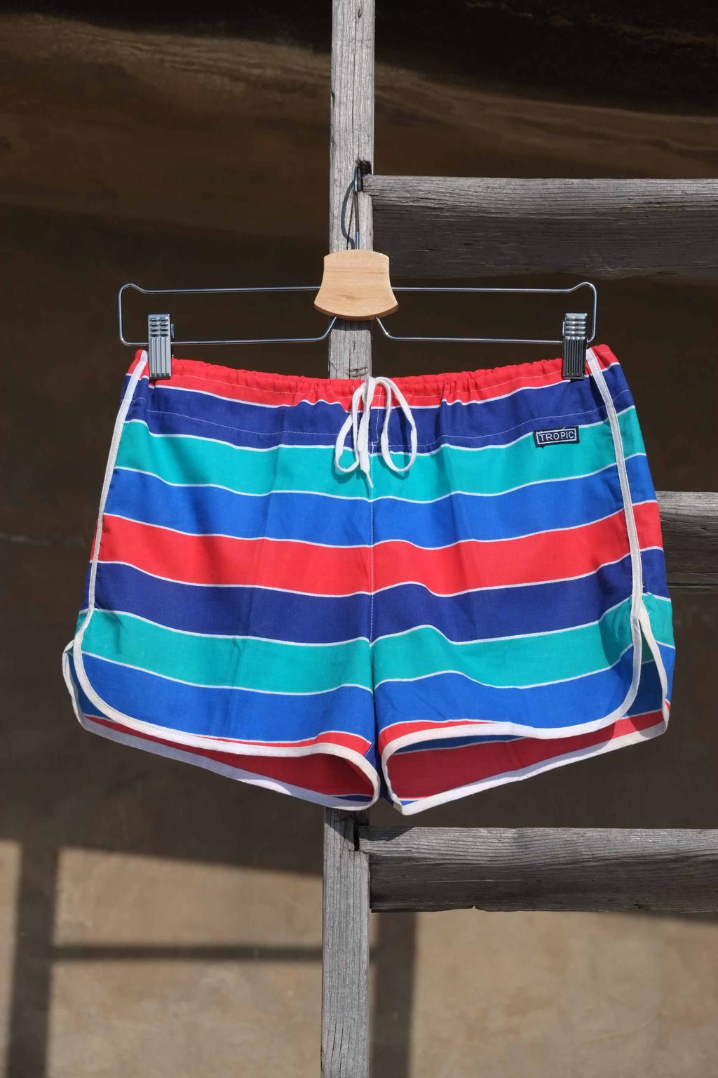 Vintage TROPIC swim shorts featuring a striped pattern in red,
 blue, green, and white. The shorts have an elastic waistband and drawstring closure. They are displayed on a hanger and placed on a wooden rack outside in the sun.