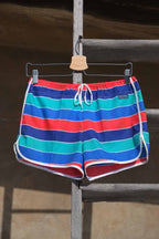 Vintage TROPIC swim shorts featuring a striped pattern in red,
 blue, green, and white. The shorts have an elastic waistband and drawstring closure. They are displayed on a hanger and placed on a wooden rack outside in the sun.