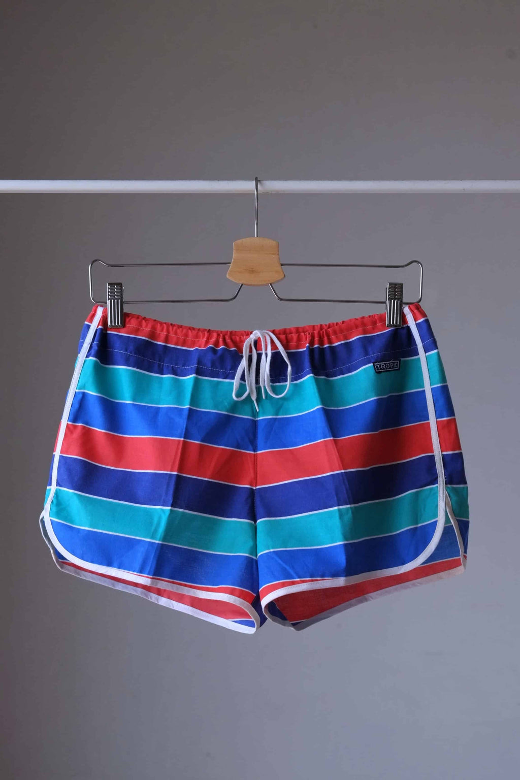Vintage TROPIC swim shorts featuring a striped pattern in red,
 blue, green, and white. The shorts have an elastic waistband and drawstring closure. Displayed on a hanger.