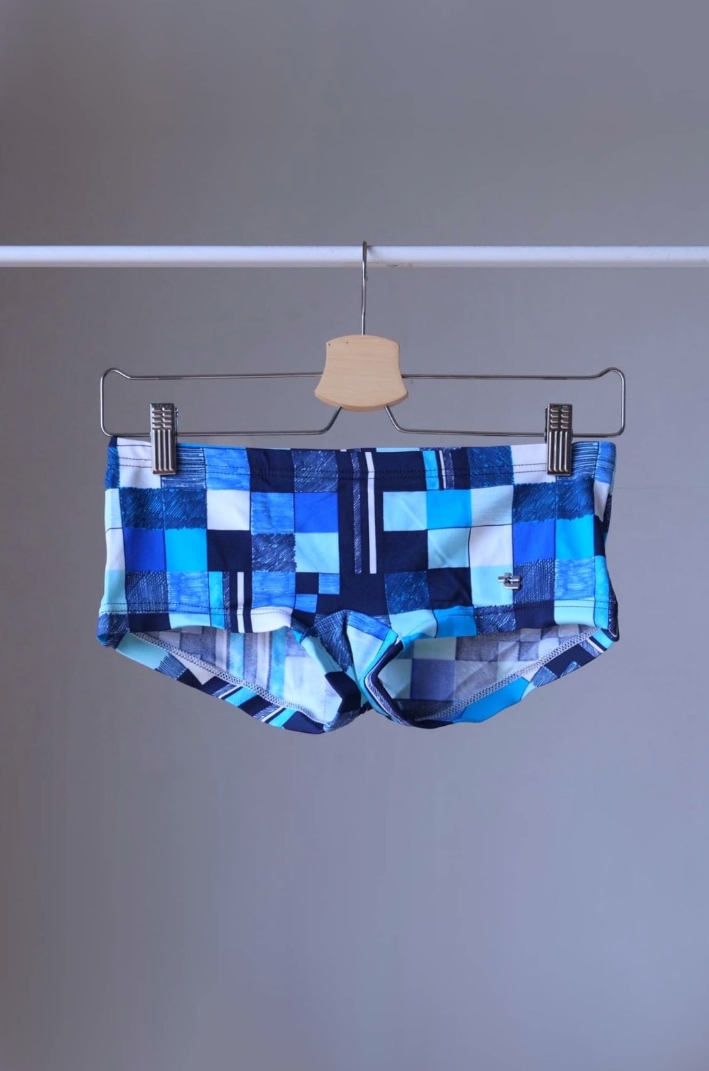 
Vintage Tropic swim briefs featuring a retro checkered pattern in shades of blue, and white. The briefs are hanging on a hanger against a gray background.