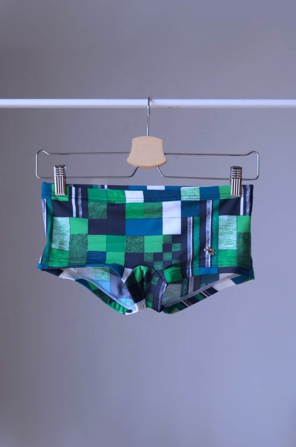 
Vintage Tropic swim briefs featuring a retro checkered pattern in shades of green and white. The briefs are hanging on a hanger against a gray background.