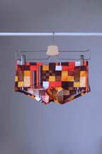 
Vintage Tropic swim briefs featuring a retro checkered pattern in orange, red, brown, and white. The briefs are hanging on a hanger against a gray background.