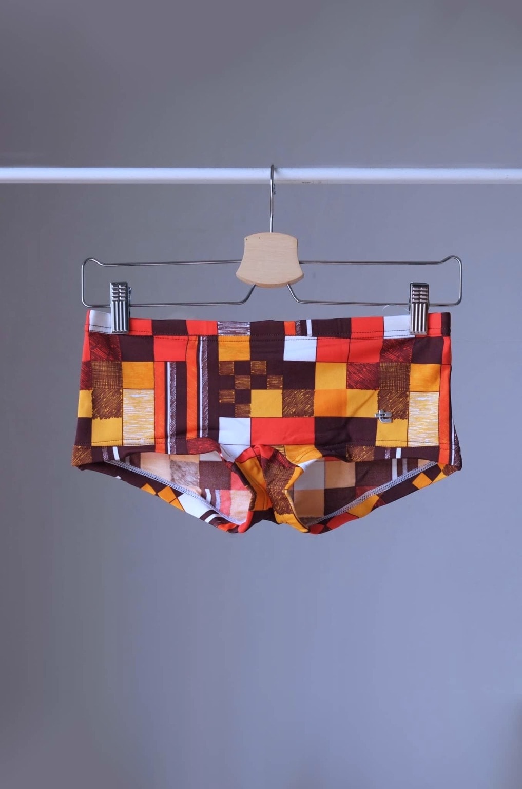 
Vintage Tropic swim briefs featuring a retro checkered pattern in orange, red, brown, and white. The briefs are hanging on a hanger against a gray background.