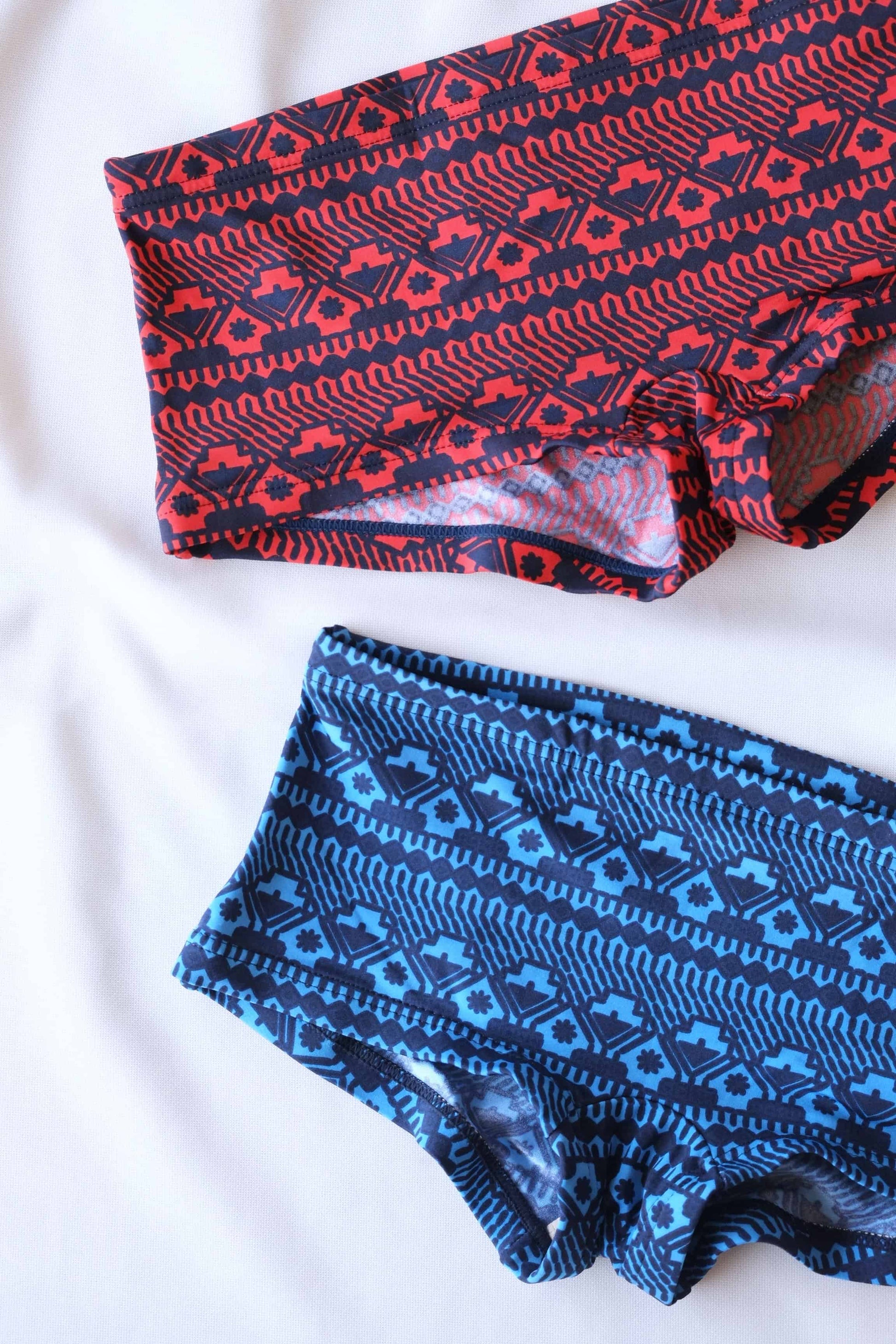 Close up of Two pairs of vintage swim briefs with a geometric pattern. One pair is primarily red with black accents, and the other pair is primarily blue with black accents. they are displayed on a white background with some palm leaves showing in the corner.