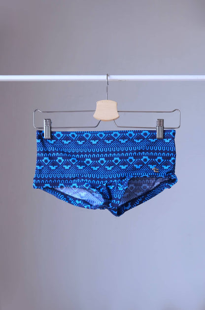 Blue swim briefs with a geometric pattern hanging on a wooden hanger. The pattern features a repeating design of triangles and squares.