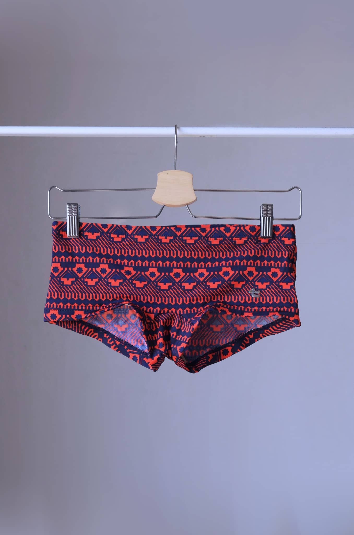 Red swim briefs with a geometric pattern hanging on a wooden hanger. The pattern features a repeating design of triangles and squares.