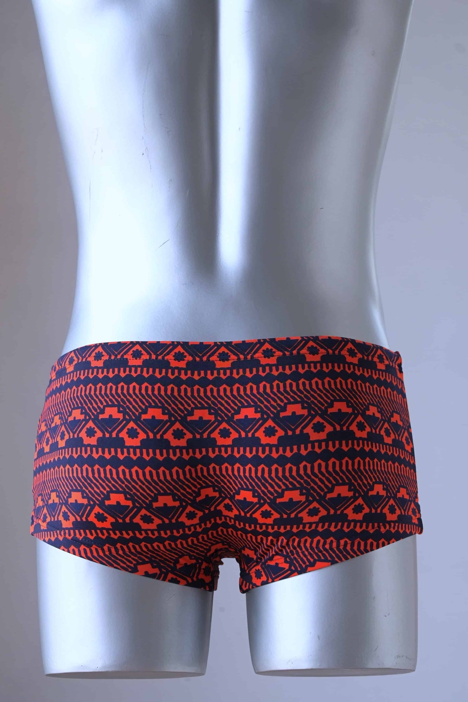 Backside of A plastic mannequin wearing a pair of vintage swim briefs with a geometric pattern primarily red with black.