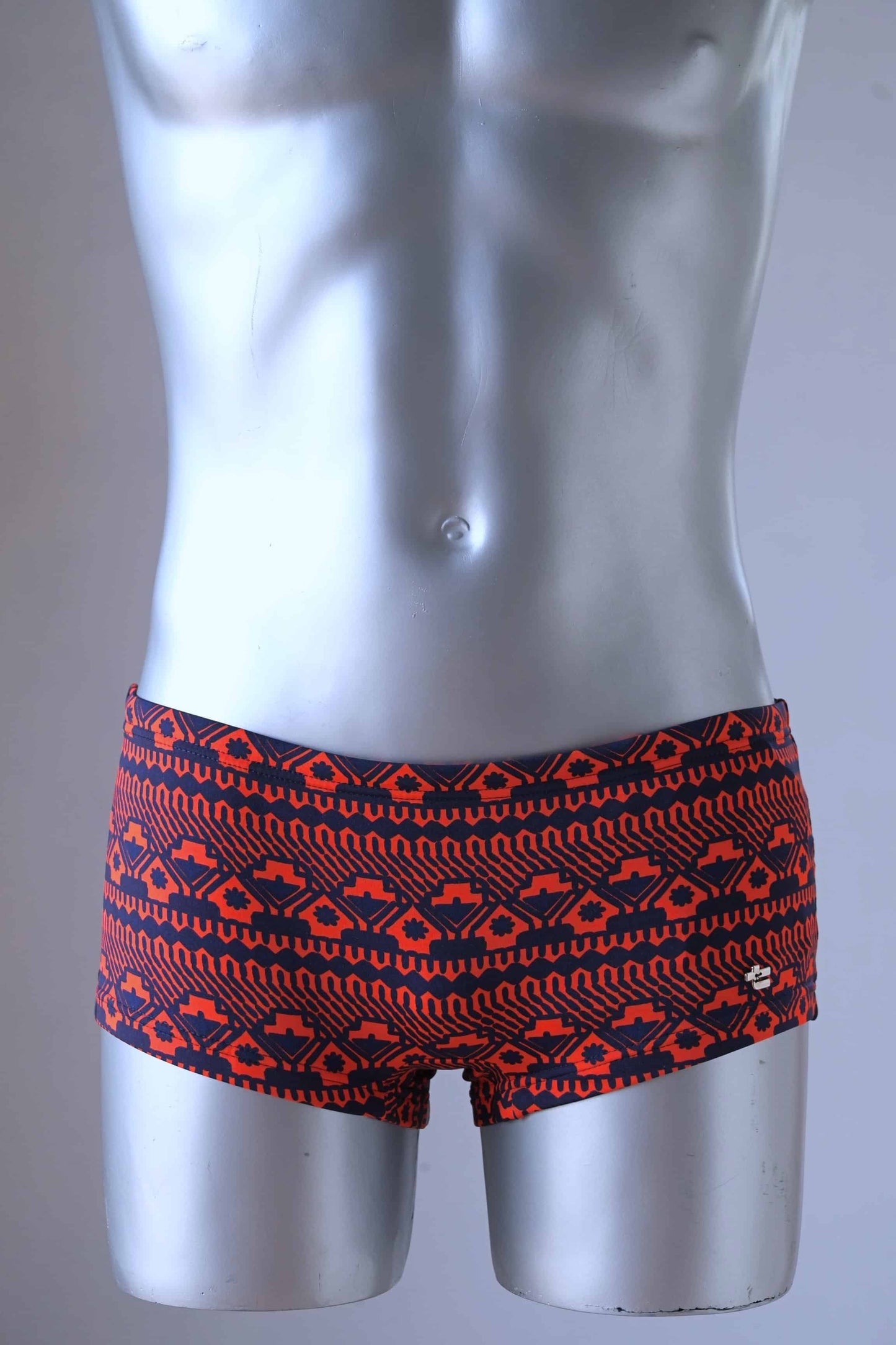 A plastic mannequin wearing a pair of vintage swim briefs with a geometric pattern primarily red with black.