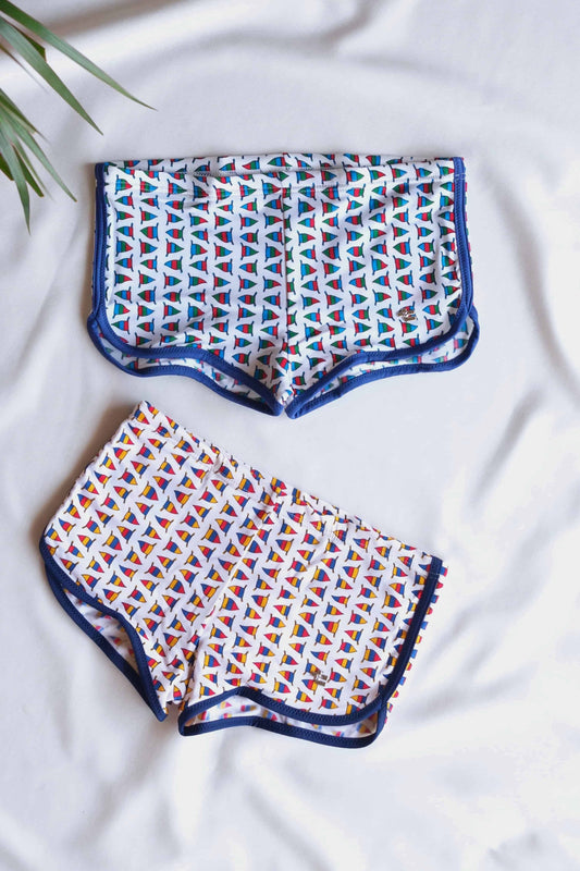 Two pairs of vintage men's swim briefs featuring a sailboat pattern.
One pair is white with navy blue trim and nautical flags, while the other pair is white with red nautical flags and navy trim. 