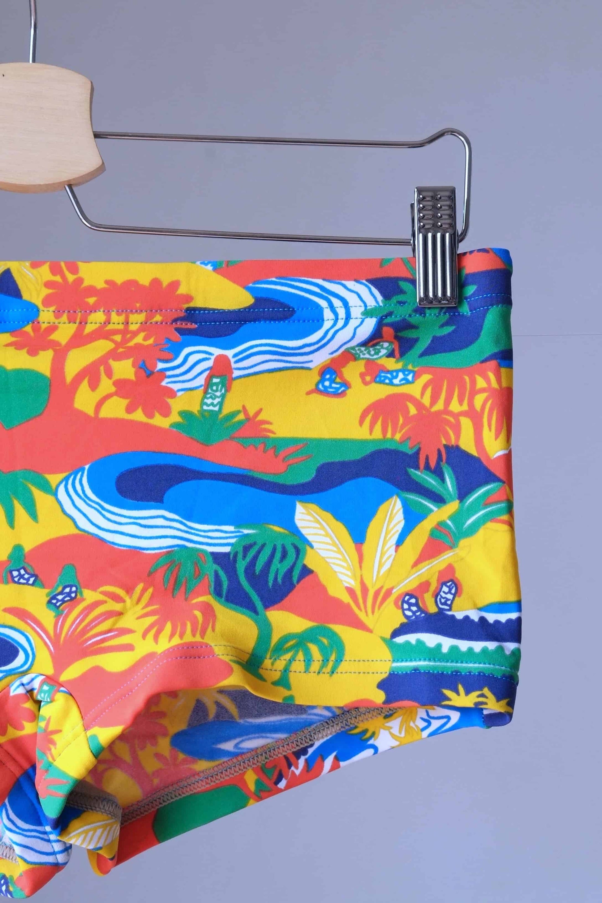 Close-up of vintage men's swim briefs featuring a tropical landscape pattern.
 The briefs are primarily orange with accents of blue, yellow, and green. The pattern includes palm trees, mountains, and water.