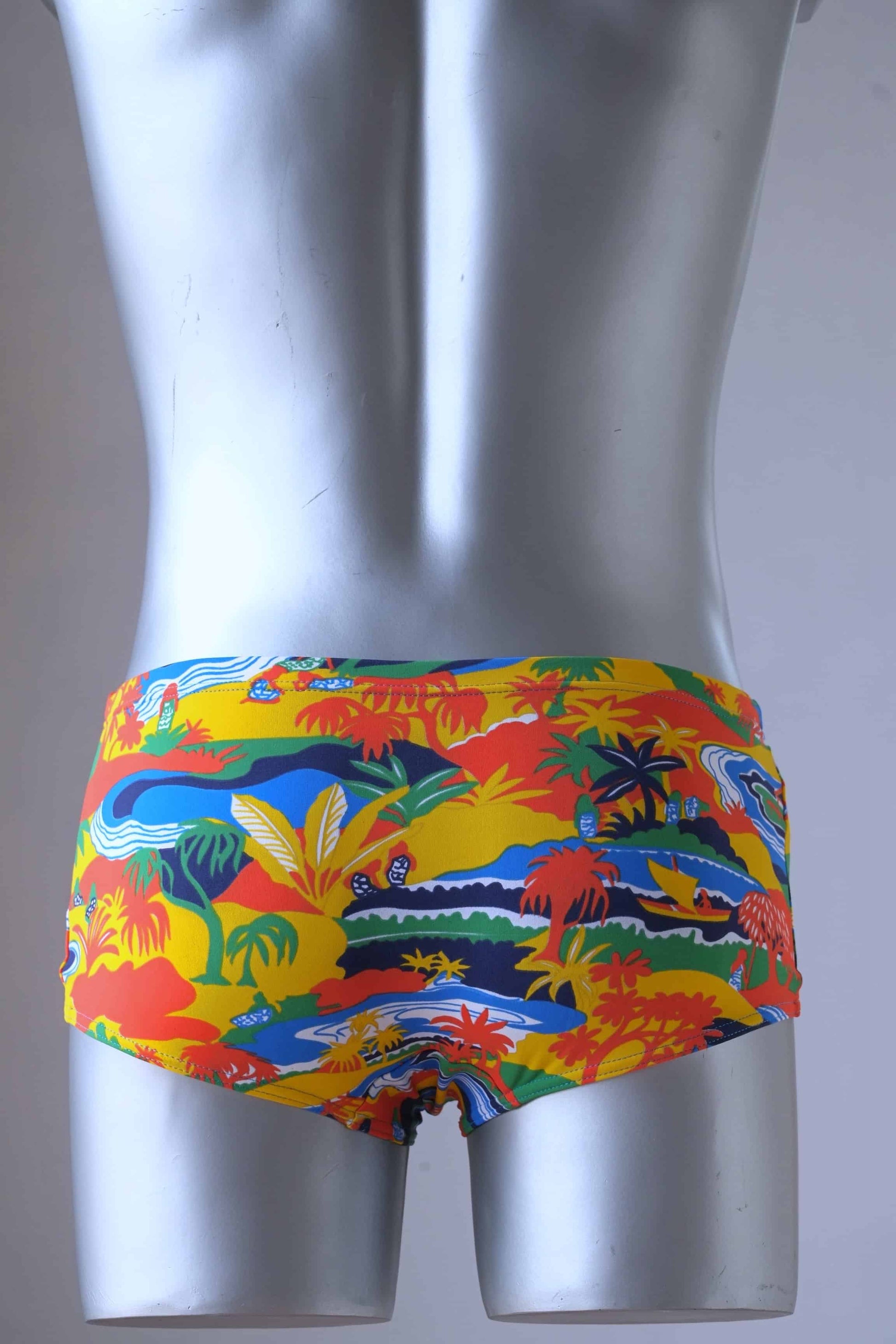 Back shot of a pair of Vintage men's swim briefs worn by a mannequin. The briefs are primarily orange and yellow with a tropical landscape pattern featuring palm trees, mountains, and water. The pattern also includes shades of blue, white, and green.