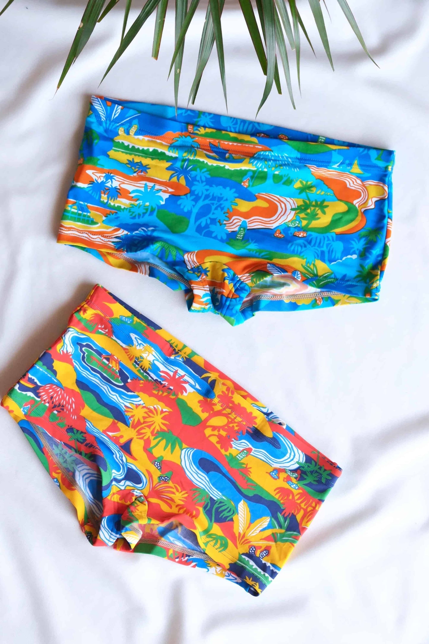 Two pairs of vintage men's swim briefs featuring a tropical landscape pattern.
 One pair is primarily blue with hints of green and yellow, while the other pair is primarily orange with hints of red and green.