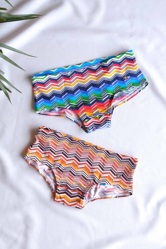 Two pairs of Tropic vintage swim briefs with a chevron pattern. One pair is primarily blue, green, and yellow. The other pair is primarily orange, red, and yellow.