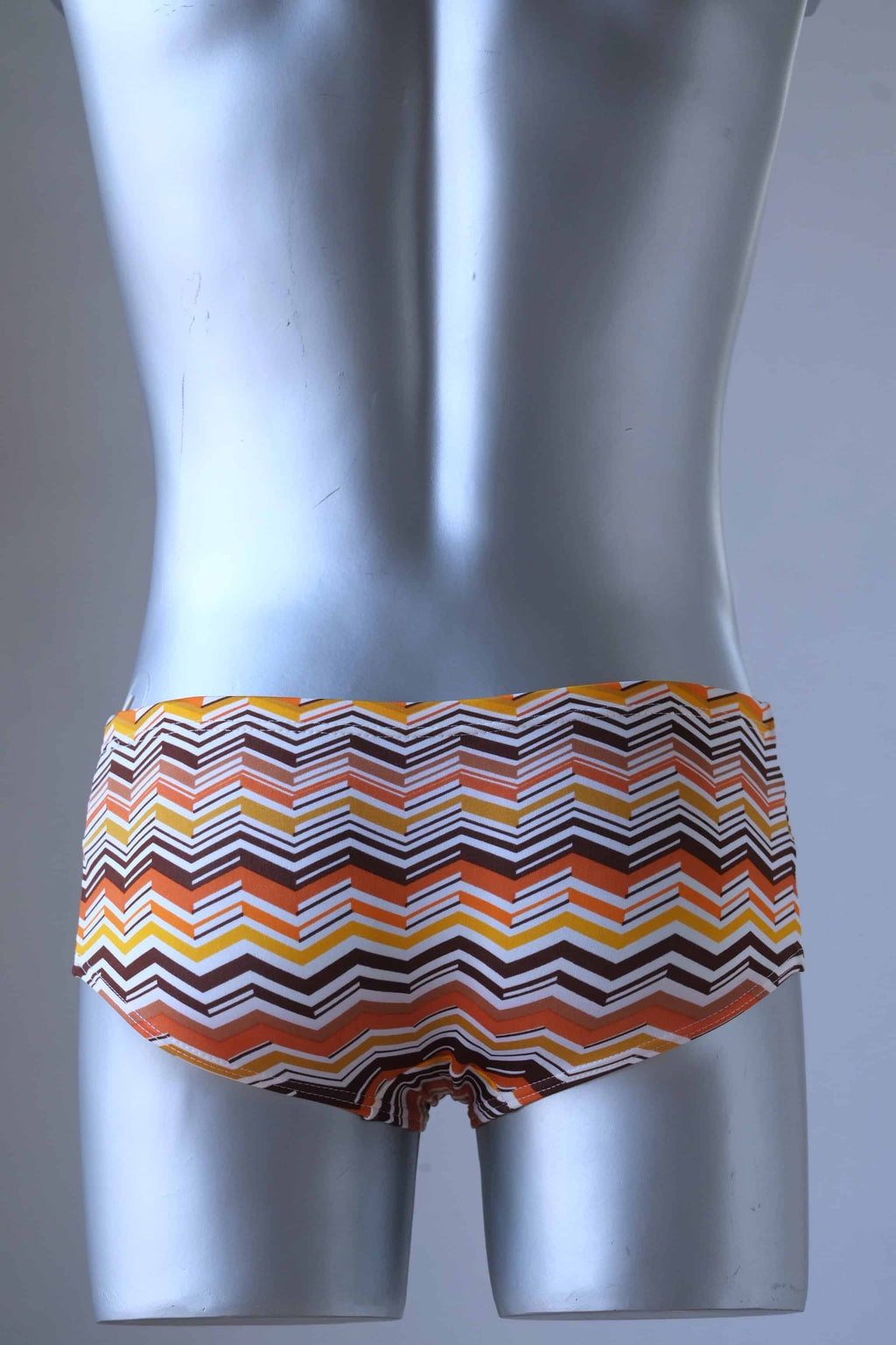 A mannequin wearing a pair of Tropic Vintage swim briefs featuring a brown, orange and yellow chevron pattern, shown from the back.