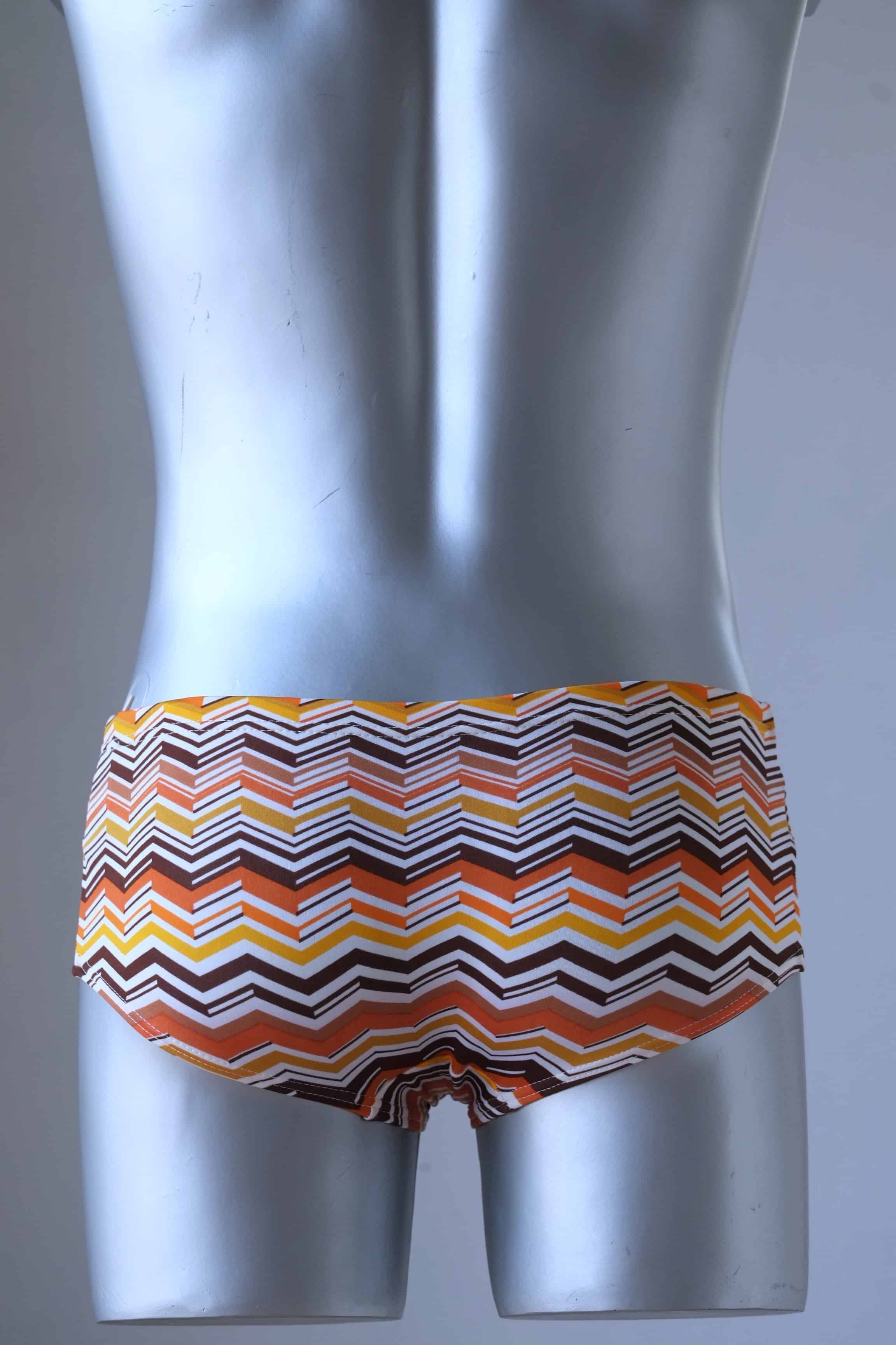 A mannequin wearing a pair of Tropic Vintage swim briefs featuring a brown, orange and yellow chevron pattern, shown from the back.