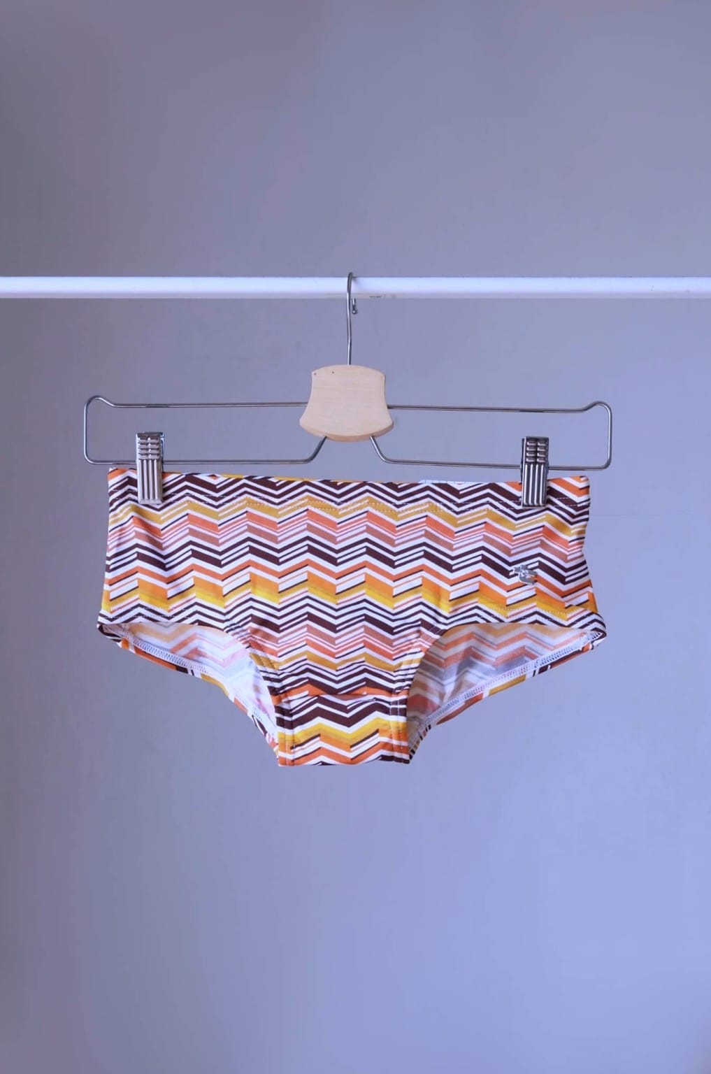 Tropic Vintage swim briefs featuring a brown, orange and yellow chevron pattern, hanging on a hanger.