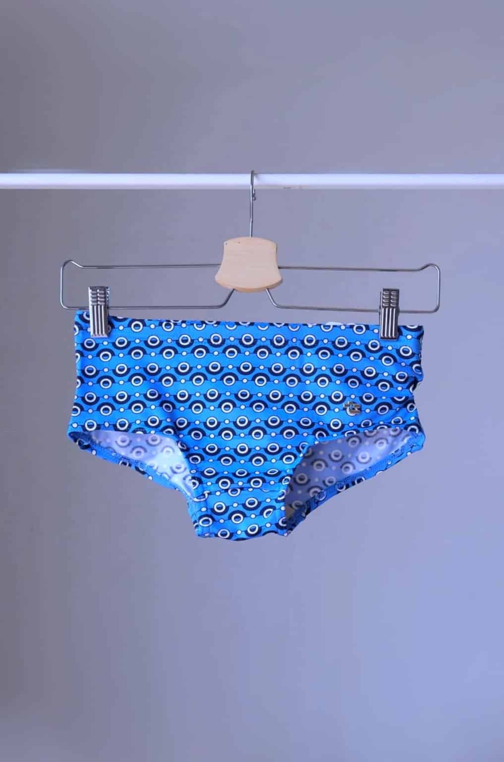 Tropic Vintage blue swim briefs with a repeating circular pattern featuring red, white, and blue circles. The briefs are hanging on a wooden hanger.