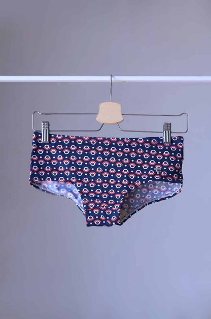 Tropic Vintage Navy and Red swim briefs with a repeating circular pattern featuring red, white, and blue circles. The briefs are hanging on a wooden hanger.