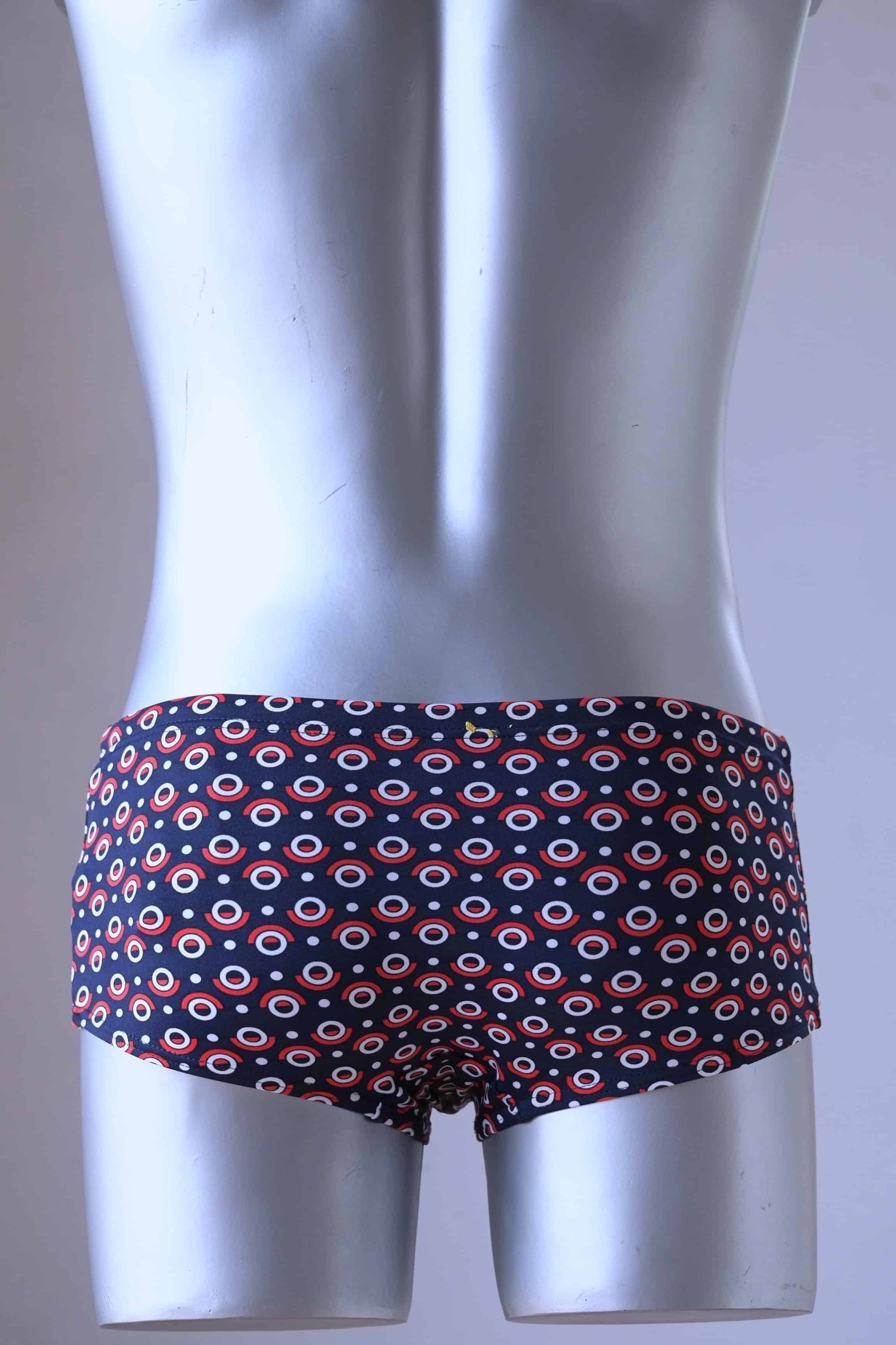 Shot from the back, A plastic mannequin wearing a pair of Tropic Vintage Navy and Red swim briefs with a repeating circular pattern featuring red, white, and blue circles.