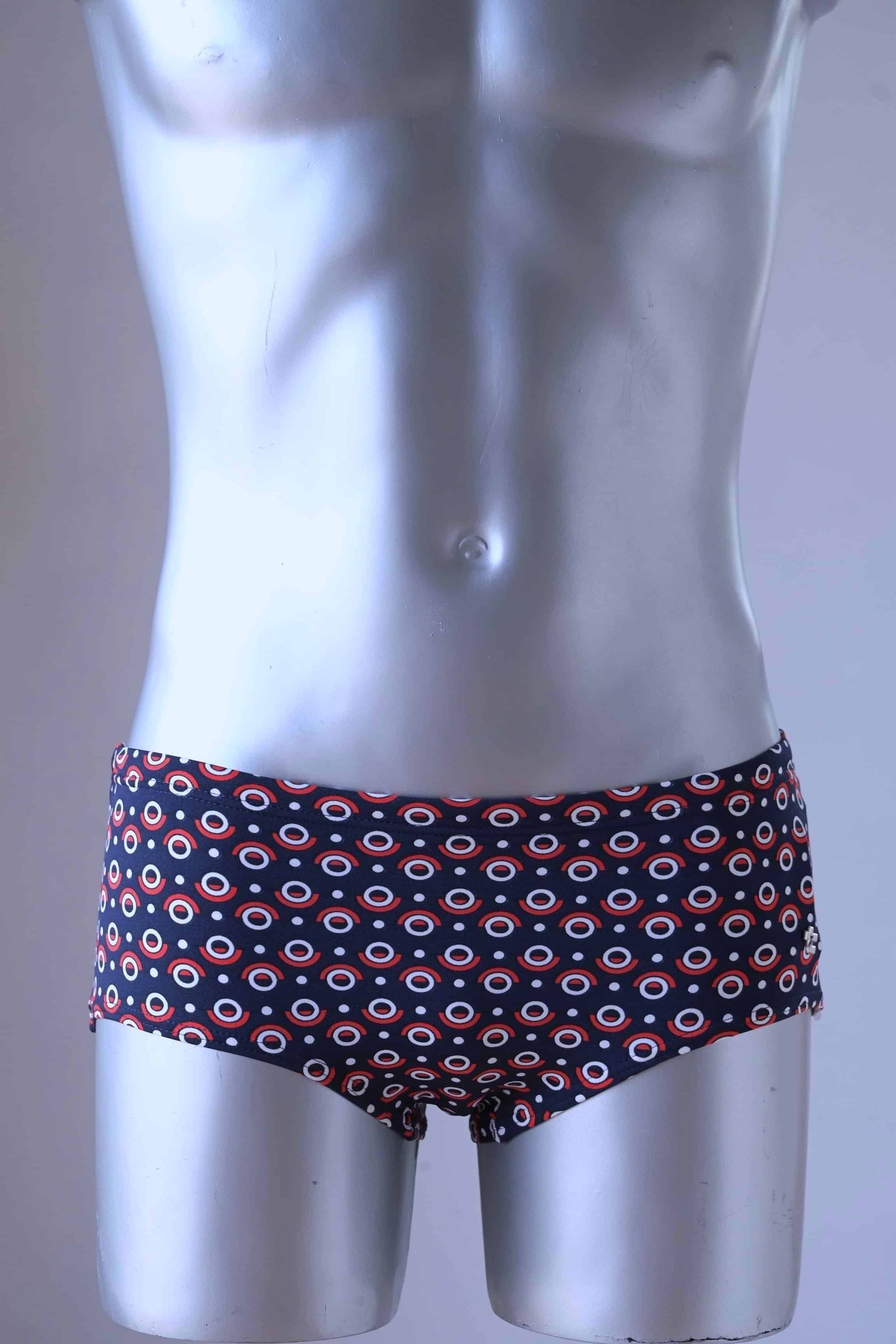 A plastic mannequin wearing a pair of Tropic Vintage Navy and Red swim briefs with a repeating circular pattern featuring red, white, and blue circles.