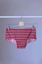 Tropic Vintage Red swim briefs with a repeating circular pattern featuring red, white, and blue circles. The briefs are hanging on a wooden hanger.