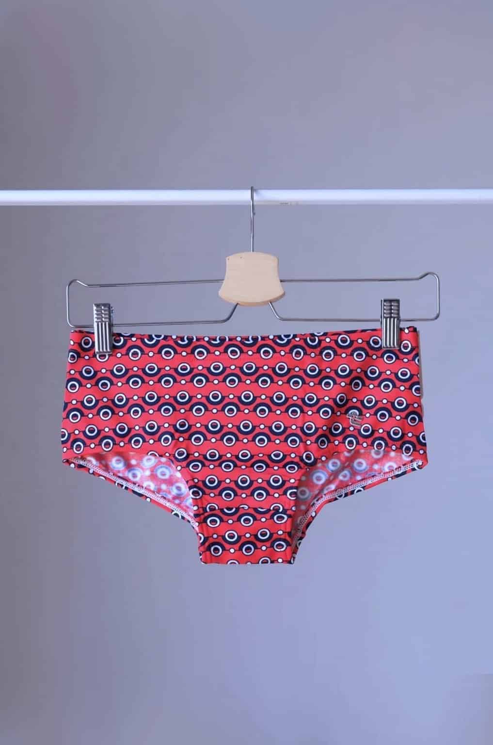 Tropic Vintage Red swim briefs with a repeating circular pattern featuring red, white, and blue circles. The briefs are hanging on a wooden hanger.